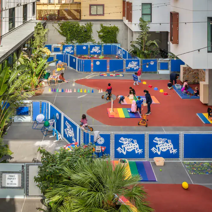 Children playing in a colorful outdoor playground with blue fencing and tropical plants.